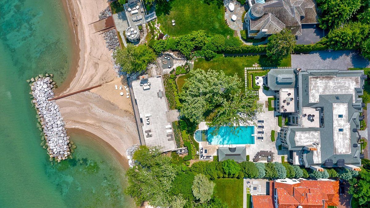 This high-angle aerial view captures a luxurious waterfront estate featuring a sprawling main residence, a private swimming pool, and an expansive stone patio. The property includes a manicured lawn, mature trees, and a private sandy beach protected by a stone breakwater extending into the clear blue water. The perspective highlights the seamless integration between the sophisticated architecture and the natural shoreline.