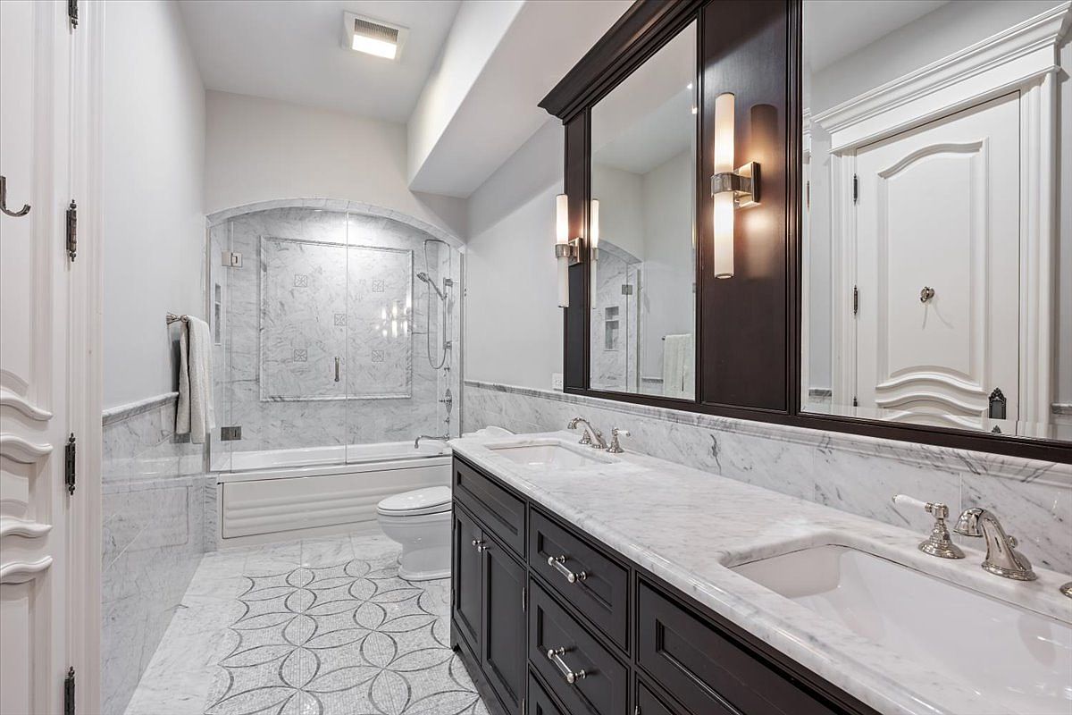 This elegant primary bathroom features a double vanity with dark cabinetry topped with light stone counters and a large mirror. A separate bathtub and shower area is enclosed by glass, showcasing intricate patterned tile flooring and stone wall accents. The space is illuminated by wall-mounted sconces and recessed lighting, creating a sophisticated and polished atmosphere.