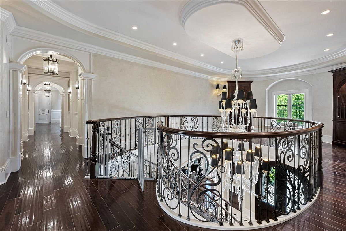 This elegant upper-level landing features dark hardwood flooring and a curved wrought-iron railing overlooking the foyer below. A grand crystal chandelier hangs from a recessed ceiling, casting light over the hallway and the intricate metalwork of the staircase. The space is characterized by a sophisticated, traditional aesthetic with arched doorways and neutral wall tones.