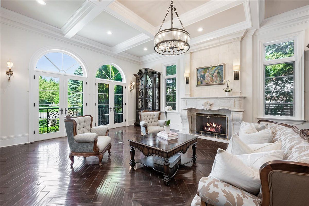 This elegant living room features dark herringbone-patterned wood flooring and a coffered ceiling with a large, multi-tiered chandelier. A prominent stone fireplace with a decorative mantel serves as the focal point, flanked by tall windows and wall sconces. The space is furnished with classic tufted armchairs and a plush sofa, creating a sophisticated and inviting atmosphere.