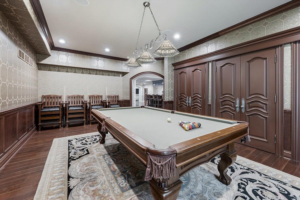 This elegant game room features a central pool table set upon a decorative area rug, surrounded by dark wood wainscoting and patterned wallpaper. A set of double doors with intricate paneling adds a formal touch, while a trio of stained-glass pendant lights hangs above the table. The perspective is centered on the pool table, looking toward the double doors and a glimpse of an adjacent bar area in the background.