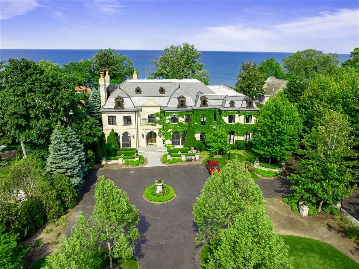 This aerial perspective captures a grand, multi-story estate featuring classic French-inspired architecture with a steep slate roof and ivy-covered stone walls. The property is situated on a lush, tree-lined lot overlooking a vast body of water, with a circular driveway and a central garden bed in the foreground. The scene conveys a sense of luxury and seclusion, highlighting the expansive grounds and the home's prominent waterfront position.