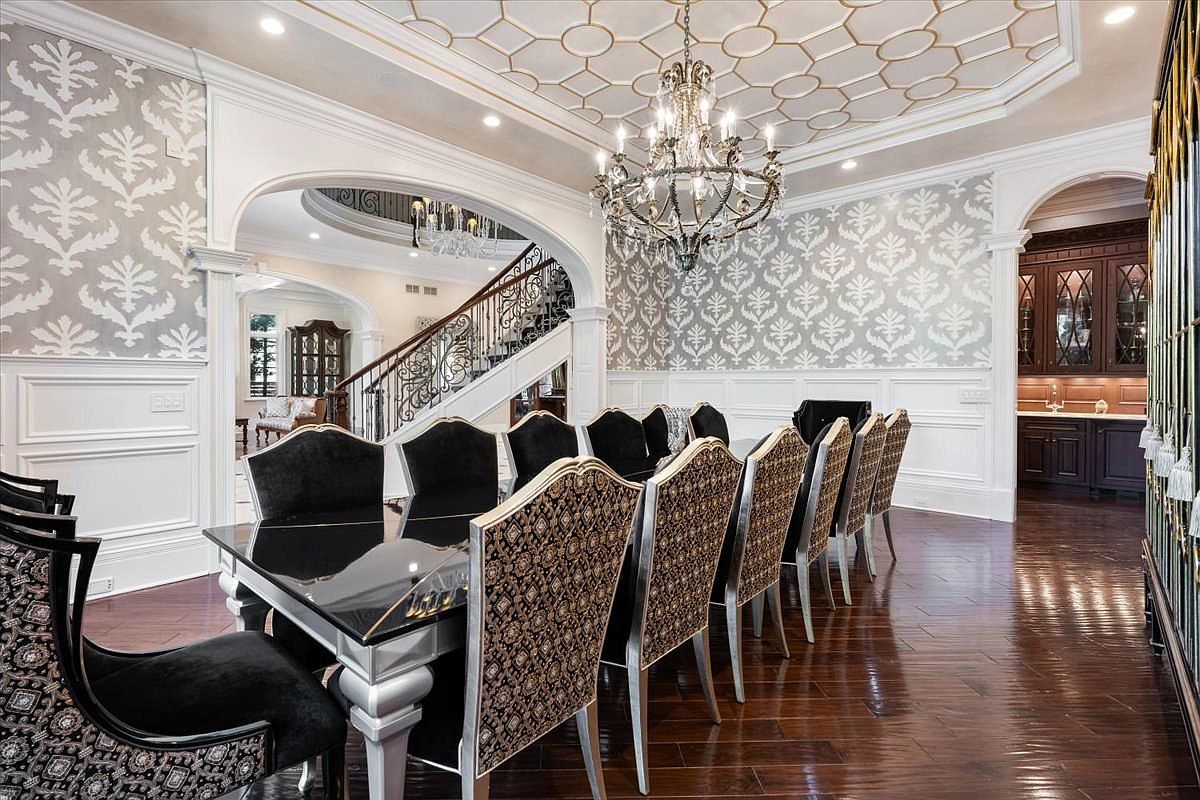 This formal dining room features a large, dark-topped table surrounded by ornate chairs with patterned upholstery and black velvet backs. The space is defined by a coffered ceiling with gold-accented geometric patterns, a crystal chandelier, and walls adorned with decorative wallpaper and wainscoting. An arched opening leads to a grand staircase, while a dark wood built-in bar area is visible in the background, creating an atmosphere of traditional luxury.
