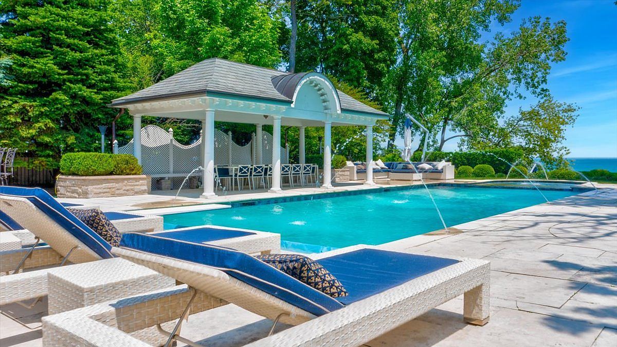 This luxurious outdoor space features a rectangular swimming pool with integrated water jets, set against a backdrop of lush green trees and a clear blue sky. A covered pavilion with white columns and a dining area provides a shaded retreat, while modern lounge chairs with blue cushions are arranged on the expansive stone patio. The scene captures a serene, resort-like atmosphere perfect for relaxation and outdoor entertaining.