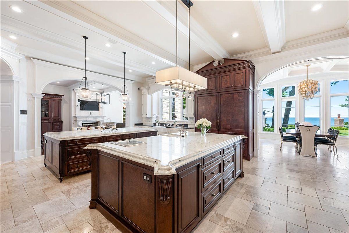 This expansive, high-end kitchen features two large islands with stone countertops and dark wood cabinetry, creating a sophisticated and symmetrical layout. The space is illuminated by elegant pendant lighting and recessed ceiling lights, while a dining area with a chandelier sits adjacent to large windows overlooking the water. The overall aesthetic is one of luxury and traditional craftsmanship, with light-toned tile flooring providing a neutral foundation for the rich wood tones.