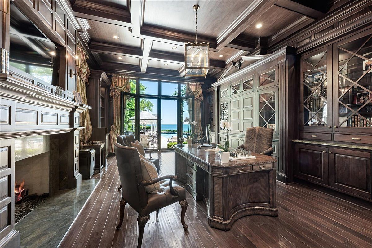 This sophisticated home office features rich, dark wood paneling, coffered ceilings, and built-in cabinetry with glass-fronted display cases. A large, ornate wooden desk sits in the center of the room, complemented by leather seating and a fireplace with a stone surround. Large windows at the rear of the room provide a clear view of an outdoor pool and the horizon beyond, creating a bright focal point in the otherwise moody, traditional space.