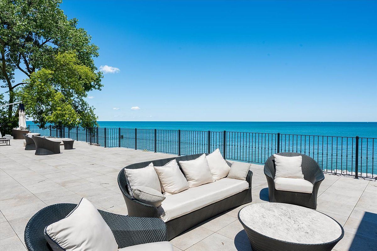 This expansive stone-paved patio offers a luxurious outdoor living space overlooking a vast, calm body of water. The area is furnished with modern, dark-wicker lounge seating featuring light-colored cushions and a matching circular stone-topped coffee table. A sleek black metal railing borders the terrace, providing an unobstructed view of the horizon under a clear blue sky.