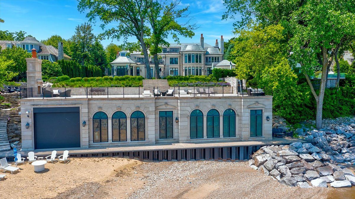 This expansive waterfront estate features a multi-level design with a prominent stone-clad boathouse at the shoreline, complete with arched windows and a large garage door. Above the boathouse, a spacious stone terrace provides an elevated outdoor living area overlooking the beach and water. The main residence sits further up the hill, showcasing classic architectural details and large windows, all set against a backdrop of lush trees and manicured landscaping.