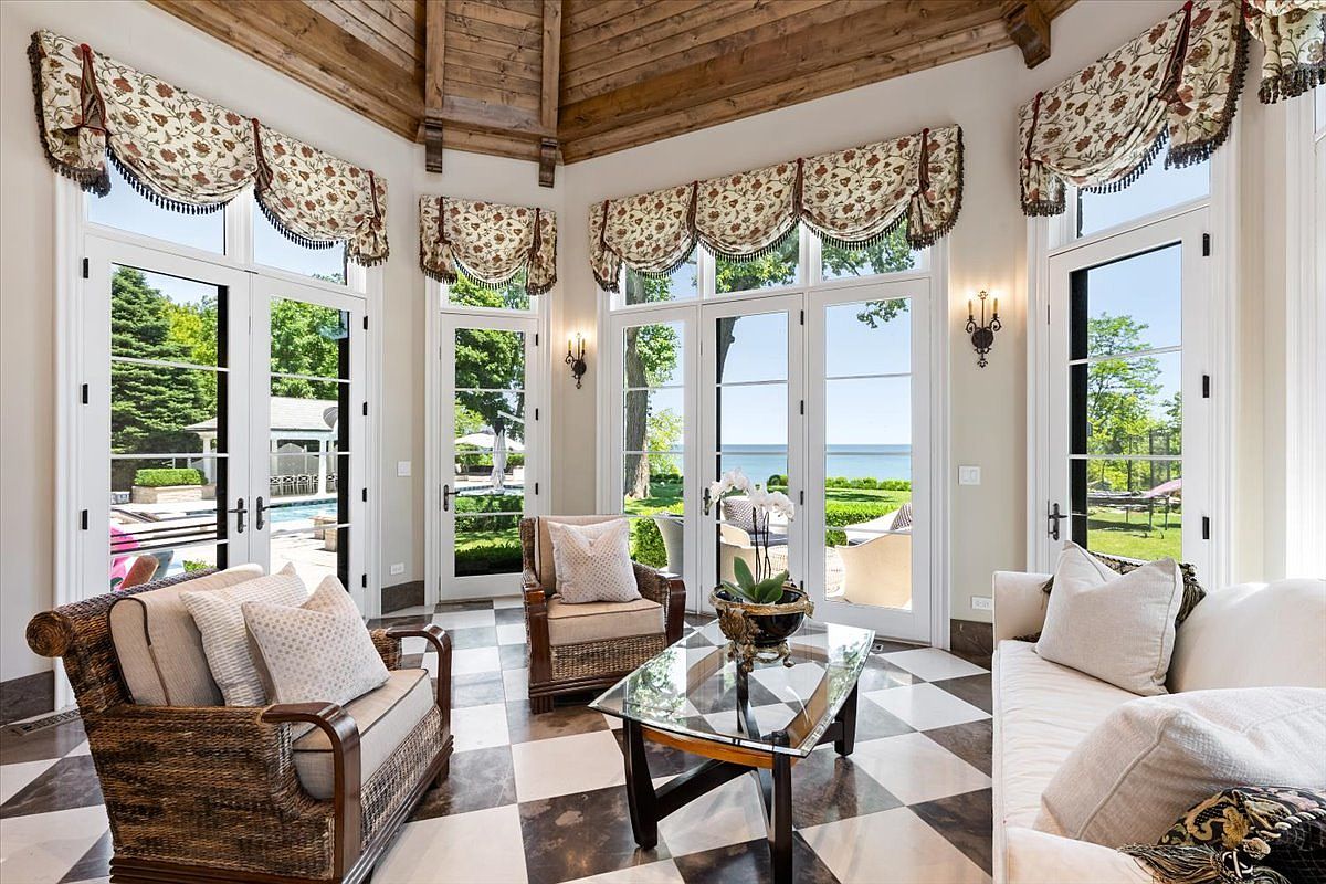 This elegant sunroom features a striking black-and-white checkered tile floor and a vaulted wood-paneled ceiling that adds warmth to the space. Multiple sets of glass doors provide abundant natural light and expansive views of the lush backyard and distant water. The room is furnished with comfortable wicker-style seating and a glass-topped coffee table, creating a serene and inviting atmosphere for relaxation.