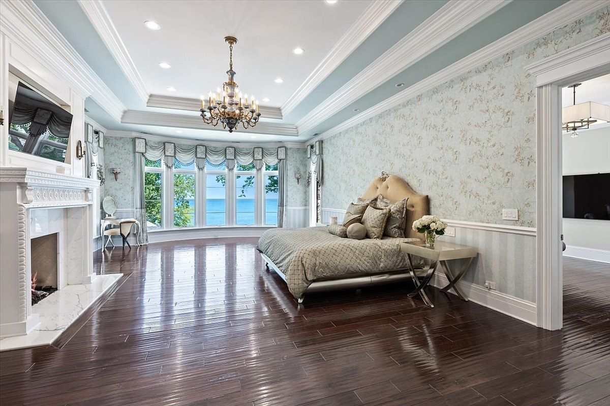 This elegant primary bedroom features a large, curved bay window offering expansive views of the water. The room is appointed with a tall, ornate fireplace surround, a crystal chandelier, and patterned wallpaper, creating a sophisticated and luxurious atmosphere. Dark, polished wood flooring extends throughout the space, which is anchored by a plush bed and a small vanity area.