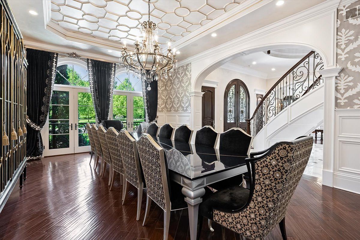 This formal dining room features a long, dark-topped table surrounded by high-backed chairs with intricate patterned upholstery. The space is defined by a dramatic crystal chandelier, a complex geometric ceiling design, and dark wood flooring that reflects the light. An arched opening leads to a staircase with ornate metal railings, while large glass doors provide a view of the lush greenery outside.
