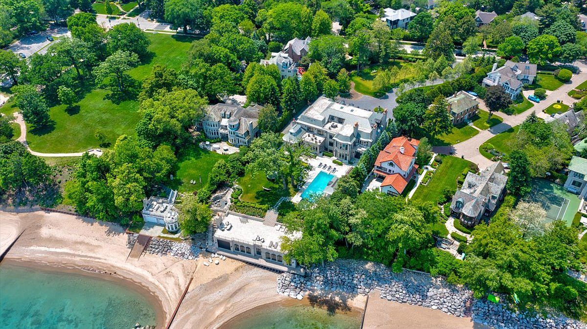 This high-angle aerial view captures a sprawling luxury estate situated directly on a sandy lakeshore. The property features a grand multi-story residence with a swimming pool, expansive manicured lawns, and a private beach-level structure, all surrounded by lush mature trees. The perspective highlights the impressive scale of the grounds and the seamless integration of the home with its waterfront environment.
