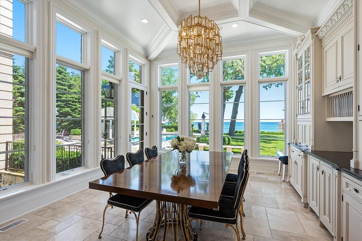 This elegant dining room features a large, polished wood table surrounded by black upholstered chairs, positioned beneath a striking gold-toned geometric chandelier. The space is defined by tall windows that offer expansive views of the lush green lawn and the blue water beyond, creating a bright and airy atmosphere. To the right, custom white cabinetry provides ample storage, while the neutral stone flooring adds a sophisticated, cohesive finish to the room.