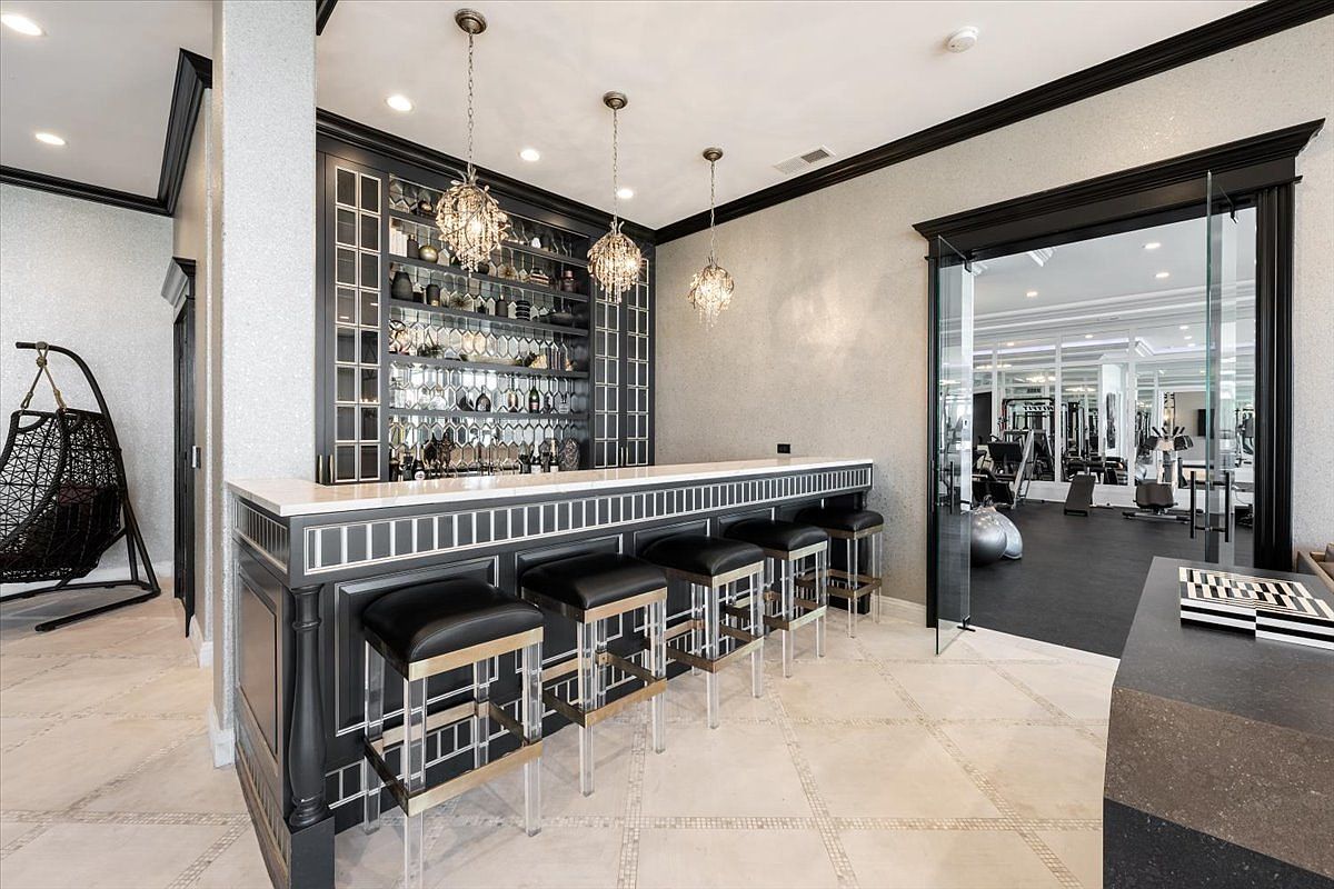 This sophisticated home bar area features a sleek, dark-toned counter with a light-colored stone top and mirrored accents, paired with four modern bar stools. Behind the bar, a built-in shelving unit displays glassware and bottles, illuminated by three elegant crystal pendant lights. To the right, a glass-paned door provides a clear view into an adjacent home gym, creating a seamless transition between entertainment and fitness spaces.
