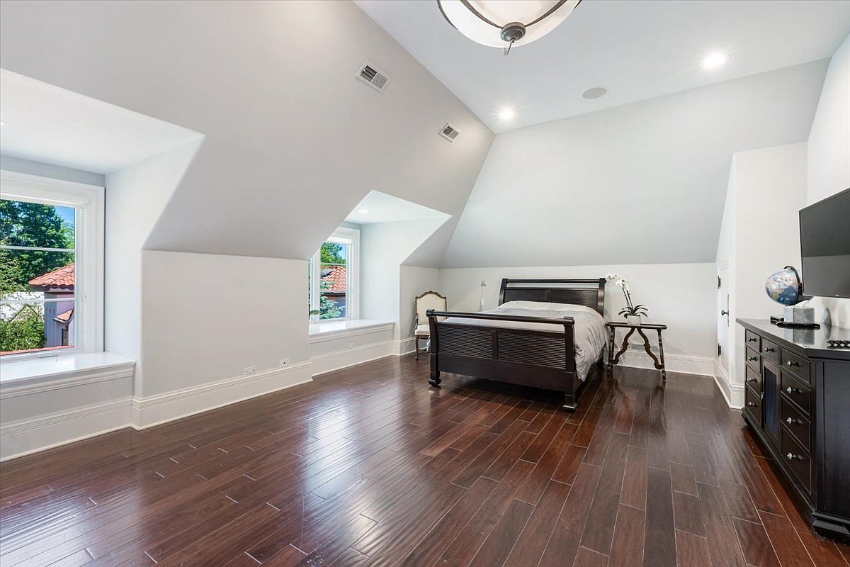 This spacious bedroom features dark wood flooring and light-colored walls, creating a clean and airy atmosphere. The room is highlighted by two dormer windows with built-in bench seating, providing natural light and a cozy nook. A dark wood bed frame and a matching dresser with a television and globe complete the furnishings, offering a balanced and functional layout.