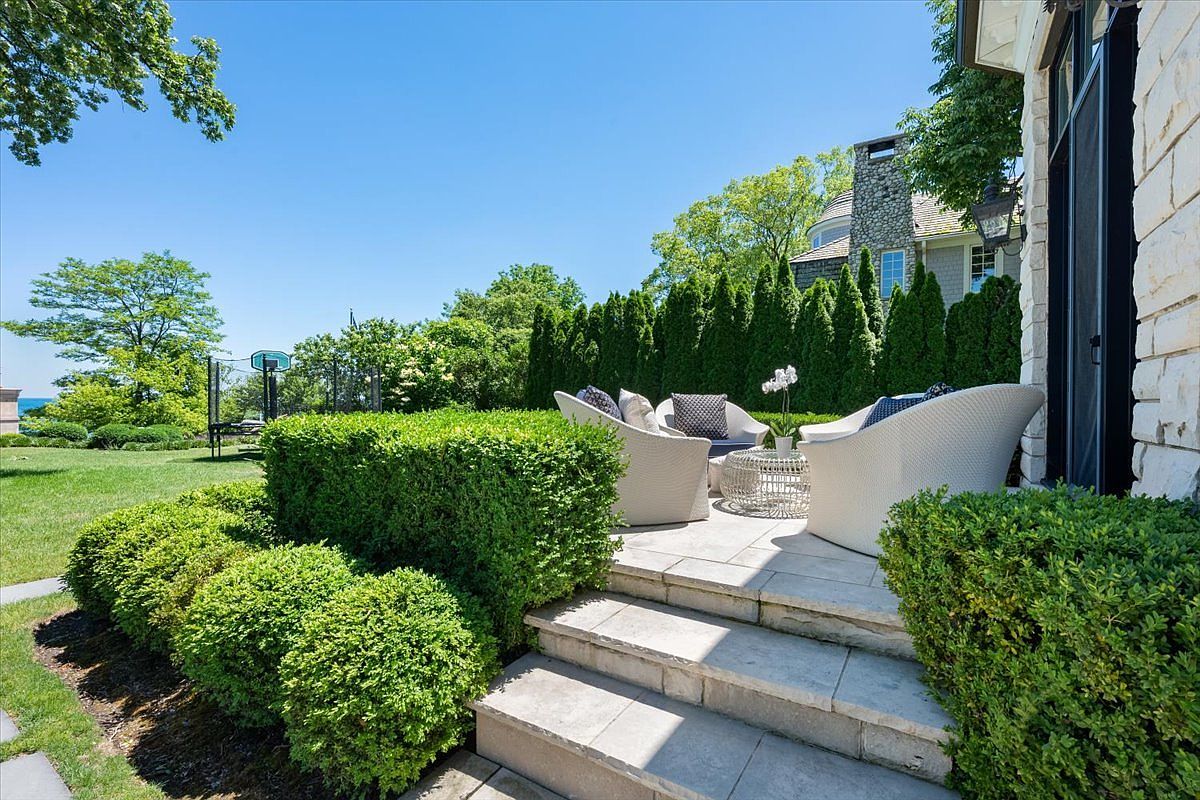 This inviting stone patio features a tiered design with comfortable white wicker seating arranged around a central fire pit. The space is framed by manicured hedges and lush greenery, creating a private outdoor retreat that overlooks a spacious lawn and distant water views. The scene is captured from a slightly elevated perspective, highlighting the seamless transition between the home's stone exterior and the landscaped backyard.