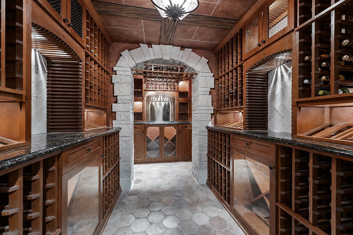 This sophisticated wine cellar features extensive custom wood racking for bottle storage, complemented by dark stone countertops and a central stone archway leading to a secondary tasting area. The space is finished with a hexagonal tile floor and a decorative ceiling fixture, creating an elegant and climate-controlled atmosphere. The perspective is centered, drawing the eye through the stone archway toward the back of the room.