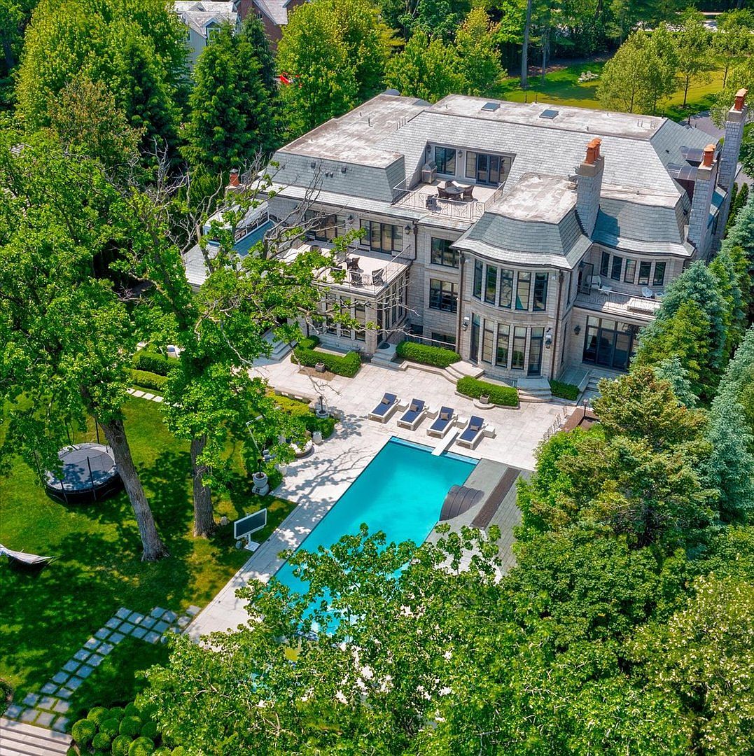 This high-angle aerial view captures a sprawling, multi-story stone estate nestled within a lush, mature landscape. The property features a large rectangular swimming pool surrounded by a stone patio, multiple terraces, and a private backyard with a trampoline and dense tree coverage. The architectural style is grand and traditional, showcasing a complex roofline with dormers and chimneys, presenting an impression of luxury and seclusion.