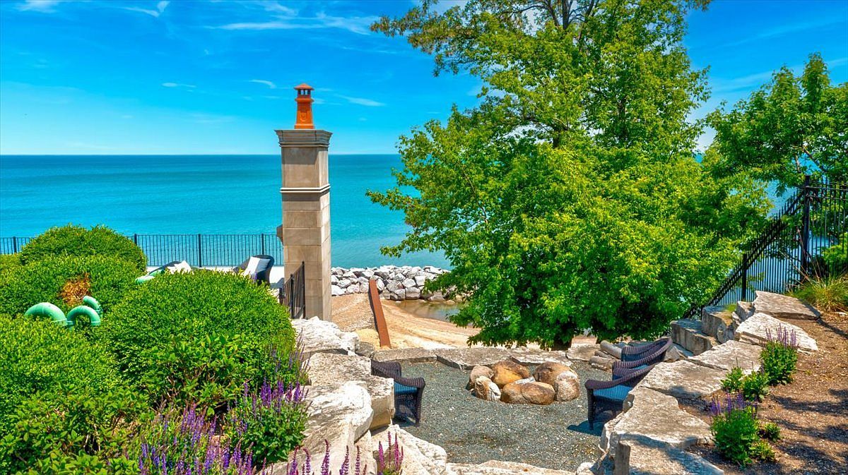 This scenic outdoor living space features a stone-paved fire pit area nestled into a tiered landscape overlooking a vast blue lake. The area includes comfortable dark wicker seating, lush green shrubbery, and a prominent stone chimney structure that adds a rustic architectural element. The perspective captures a serene, elevated view of the shoreline, emphasizing the property's prime waterfront location and tranquil atmosphere.