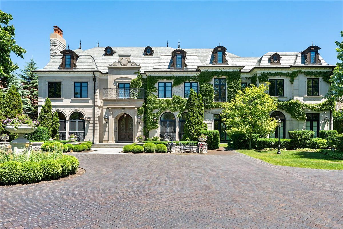 This grand, multi-story estate features a classic European-inspired architectural style with a symmetrical facade, stone exterior, and a steep slate roof punctuated by dormer windows. Lush climbing ivy covers sections of the stone walls, while a meticulously manicured landscape and a large, circular brick-paved driveway create an impressive and welcoming entrance. The perspective is a straight-on, eye-level shot that captures the full scale and stately presence of the residence.