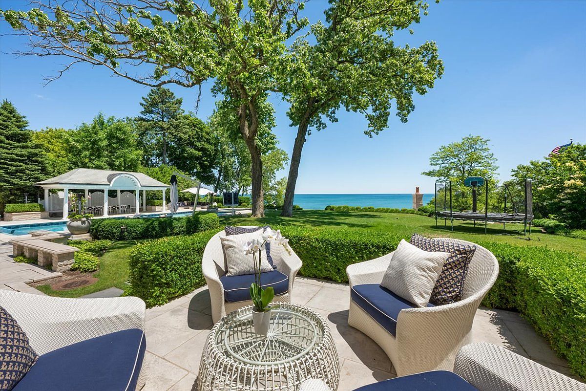 This inviting outdoor patio space features a circular wicker seating arrangement with navy blue cushions, centered around a matching glass-topped table. The patio overlooks a lush, manicured lawn that leads toward a scenic waterfront view, complemented by a nearby swimming pool and a classic gazebo. The perspective captures a serene, resort-like backyard setting, framed by mature trees and vibrant green landscaping under a clear blue sky.