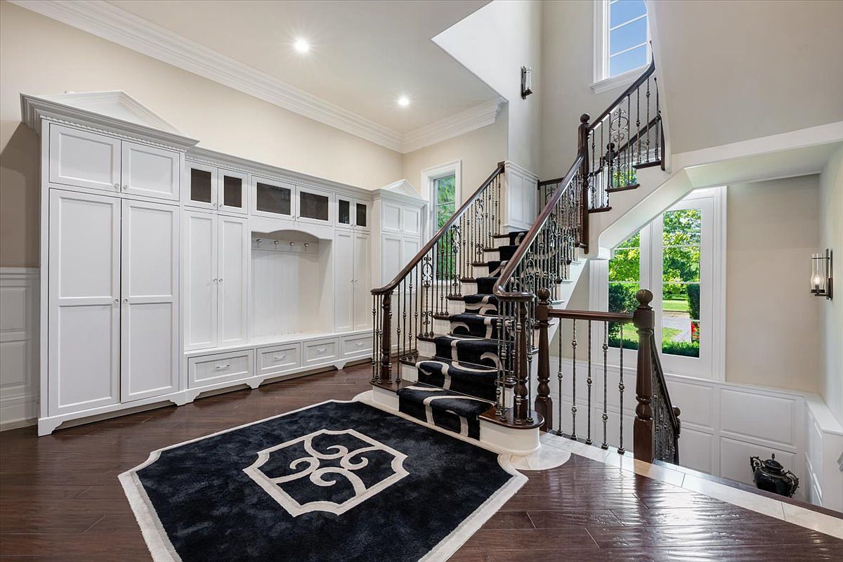 This elegant foyer features a grand staircase with dark wood railings and a striking black-and-white patterned runner. To the left, a large, custom-built white storage unit provides ample organization with integrated cabinetry and a bench area. The space is finished with dark wood flooring, a matching patterned area rug, and tall windows that allow natural light to brighten the entryway.