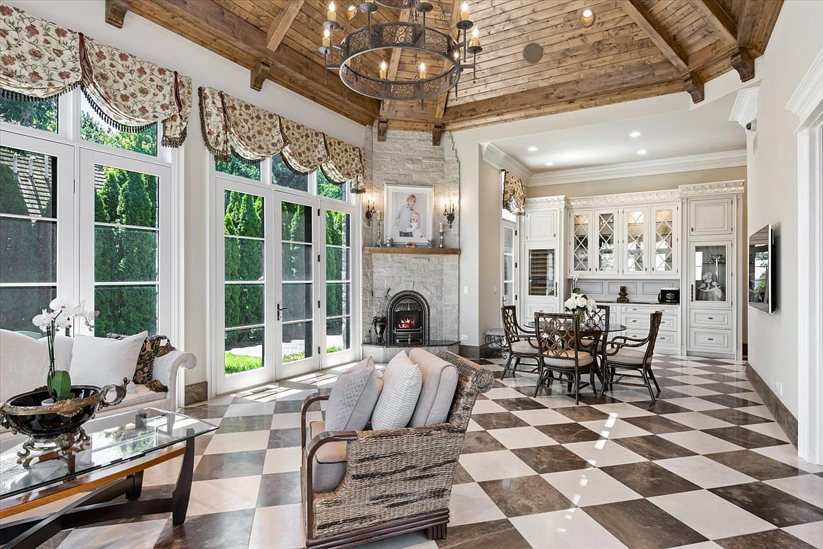 This elegant living area features a dramatic vaulted ceiling with exposed wood beams and a large iron chandelier. A stone-clad chimney breast serves as a focal point, complemented by a checkered tile floor and expansive windows draped in patterned valances. The space is furnished with comfortable seating and a small dining set, creating a sophisticated and inviting atmosphere.