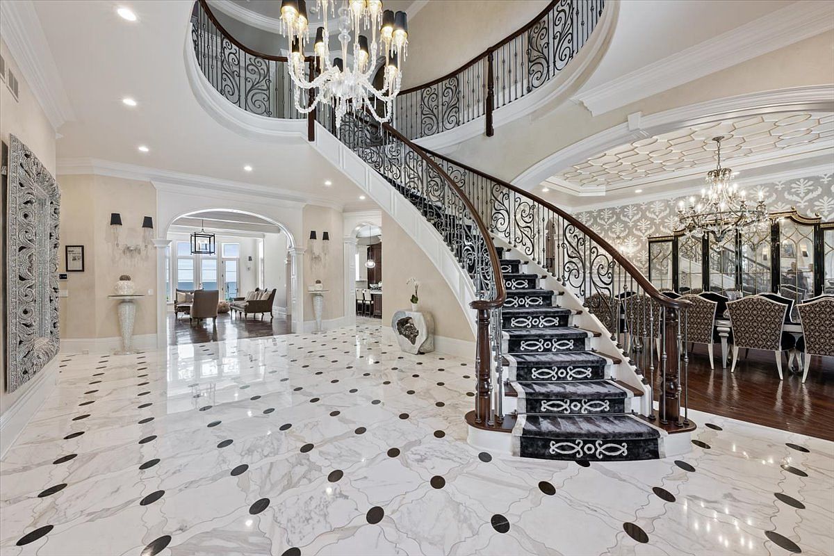 This grand foyer features a sweeping curved staircase with intricate wrought-iron railings and a patterned runner. The space is anchored by a polished stone floor with a repeating circular inlay pattern and is illuminated by a large, multi-tiered crystal chandelier. To the right, a formal dining area is visible, while an arched opening leads to a bright living space in the background, creating an open and luxurious atmosphere.