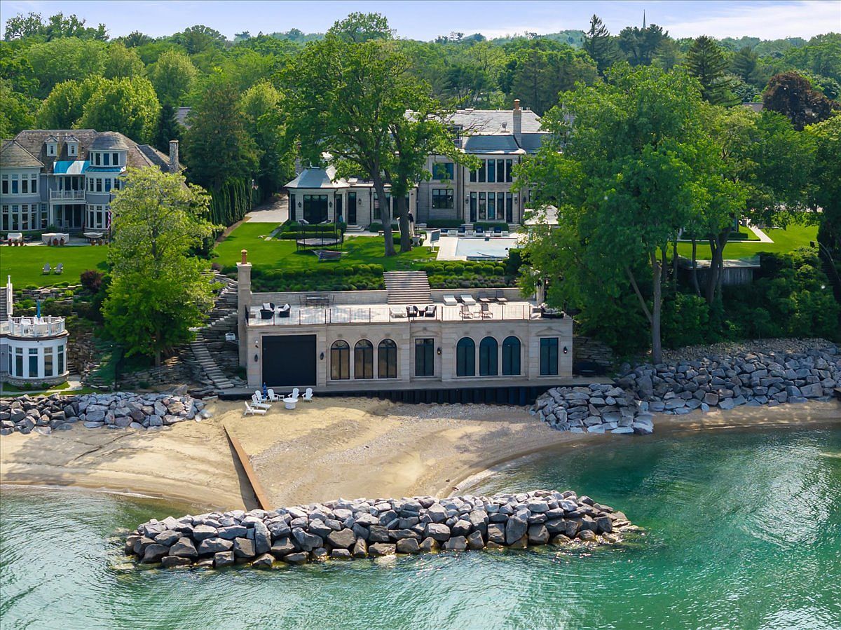 This aerial perspective captures a luxurious waterfront estate featuring a grand multi-story residence, a private swimming pool, and a sophisticated stone-clad boathouse situated directly on the beach. The property boasts expansive manicured lawns, mature trees, and a private stone breakwater protecting the sandy shoreline. The scene conveys an atmosphere of exclusivity and high-end coastal living, showcasing the seamless integration between the architecture and the natural landscape.