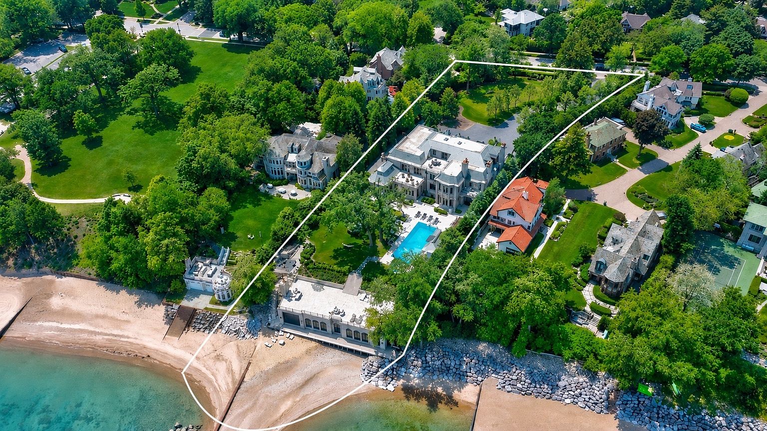 This high-angle aerial view captures a sprawling luxury estate situated directly on a sandy lakeshore. The property features a grand main residence, a private swimming pool, and a secondary structure positioned right at the water's edge, all enclosed within a highlighted boundary. Lush green lawns, mature trees, and neighboring upscale homes complete this prestigious waterfront setting.