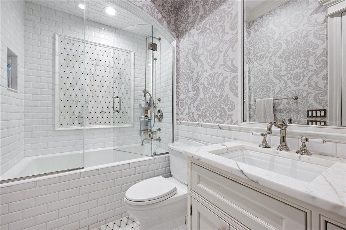 This elegant bathroom features a combination tub and shower enclosure with white subway tile walls and a decorative patterned tile accent wall. A white vanity with a stone countertop and polished metal fixtures sits beside a toilet, while the walls are adorned with a sophisticated damask-patterned wallpaper. The perspective captures the entire functional layout of the room, highlighting the blend of classic textures and clean, bright finishes.