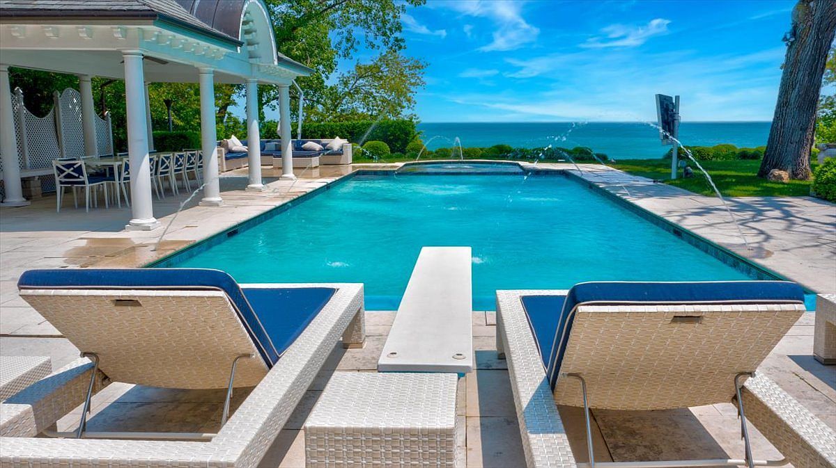 This luxurious outdoor space features a rectangular swimming pool with integrated fountains, set against a backdrop of a vast blue lake and clear sky. In the foreground, two white lounge chairs with navy blue cushions sit on a stone patio, while a covered pavilion with white columns provides a shaded seating area to the left. The scene captures a serene, high-end resort-style atmosphere perfect for relaxation and entertaining.