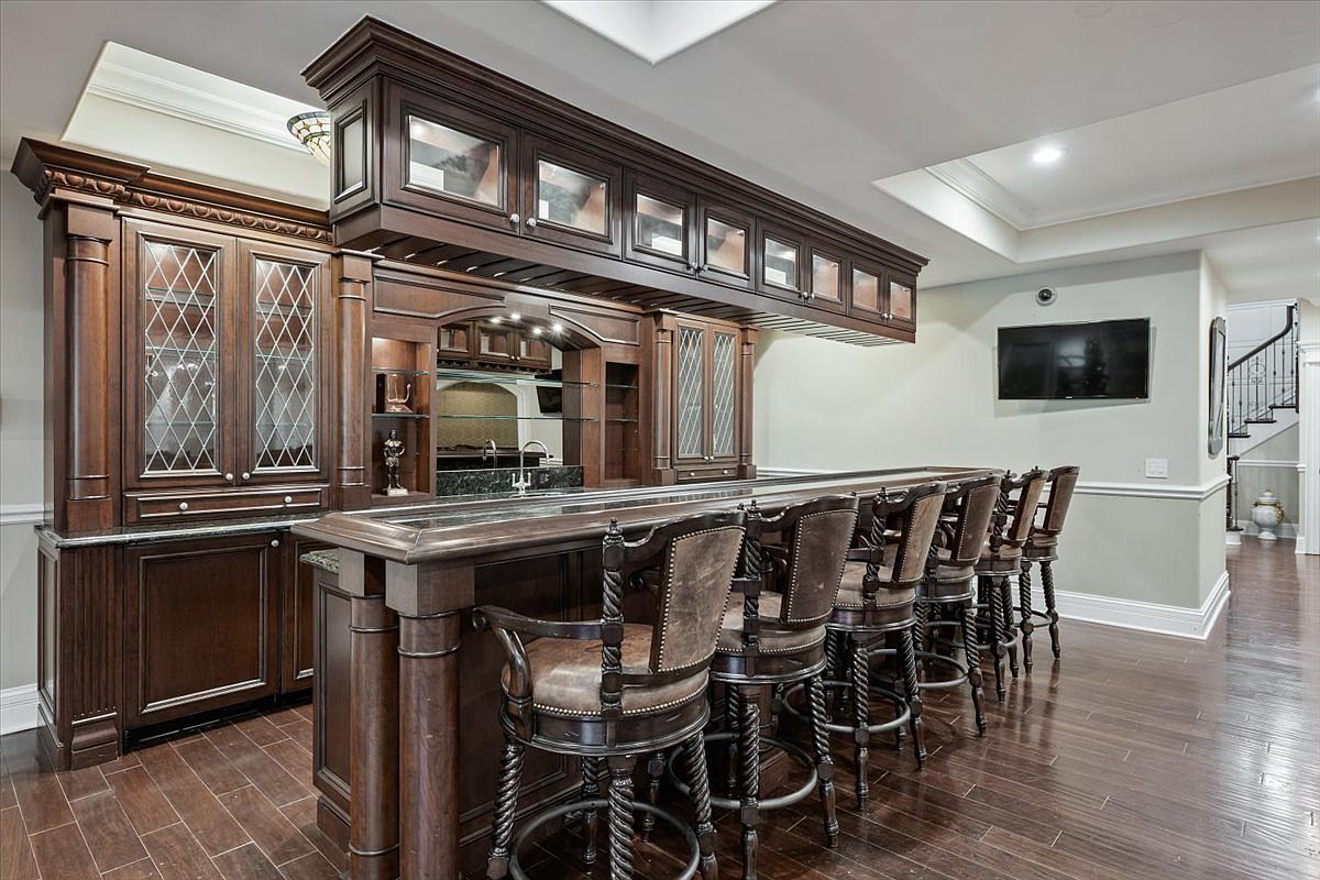 This sophisticated home bar area features rich, dark wood cabinetry with glass-fronted upper cabinets and an integrated wine rack. A long, polished bar counter is lined with several high-backed, upholstered bar stools, creating an inviting space for entertaining. The room is finished with dark wood flooring and recessed lighting, offering a classic and upscale aesthetic.