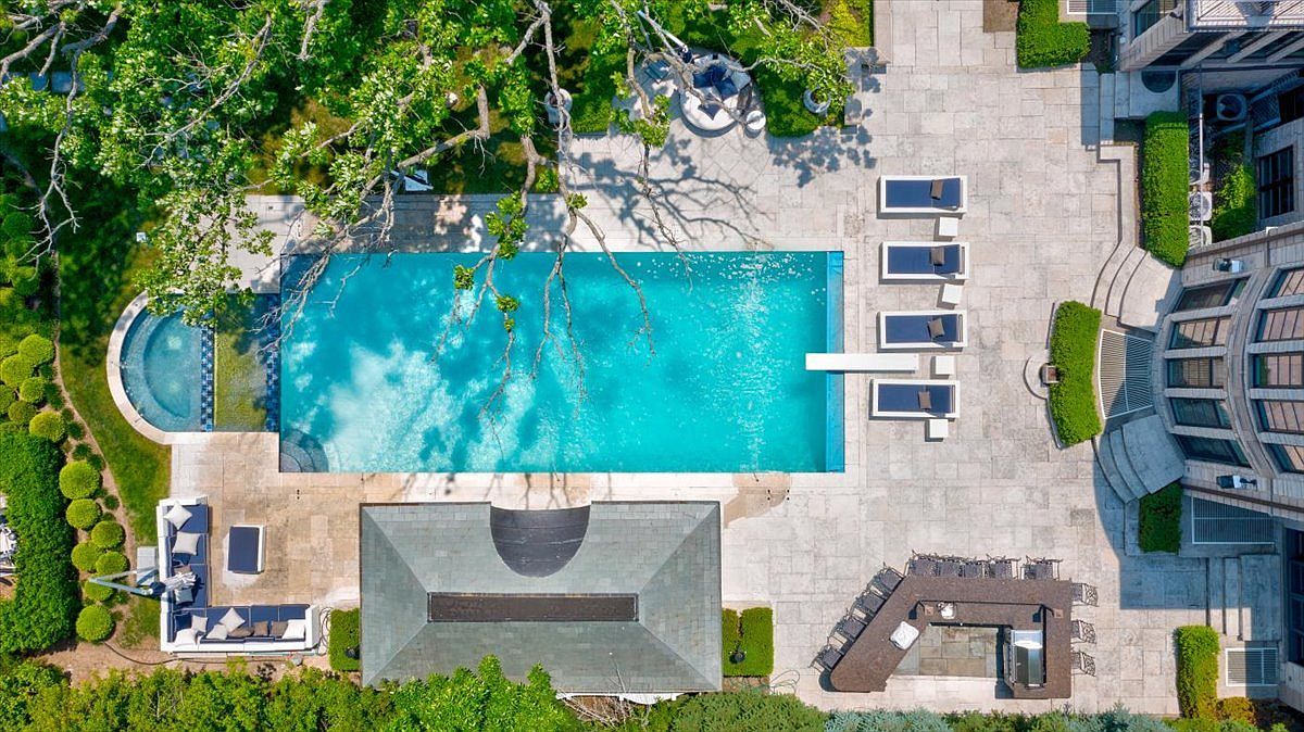 This high-angle aerial view captures a luxurious backyard oasis featuring a large rectangular swimming pool with an integrated spa. The expansive stone patio is furnished with multiple lounge chairs, a covered outdoor kitchen area, and a separate seating lounge, all surrounded by manicured hedges and mature trees. The perspective provides a comprehensive look at the property's outdoor entertainment layout and its seamless integration with the main residence.