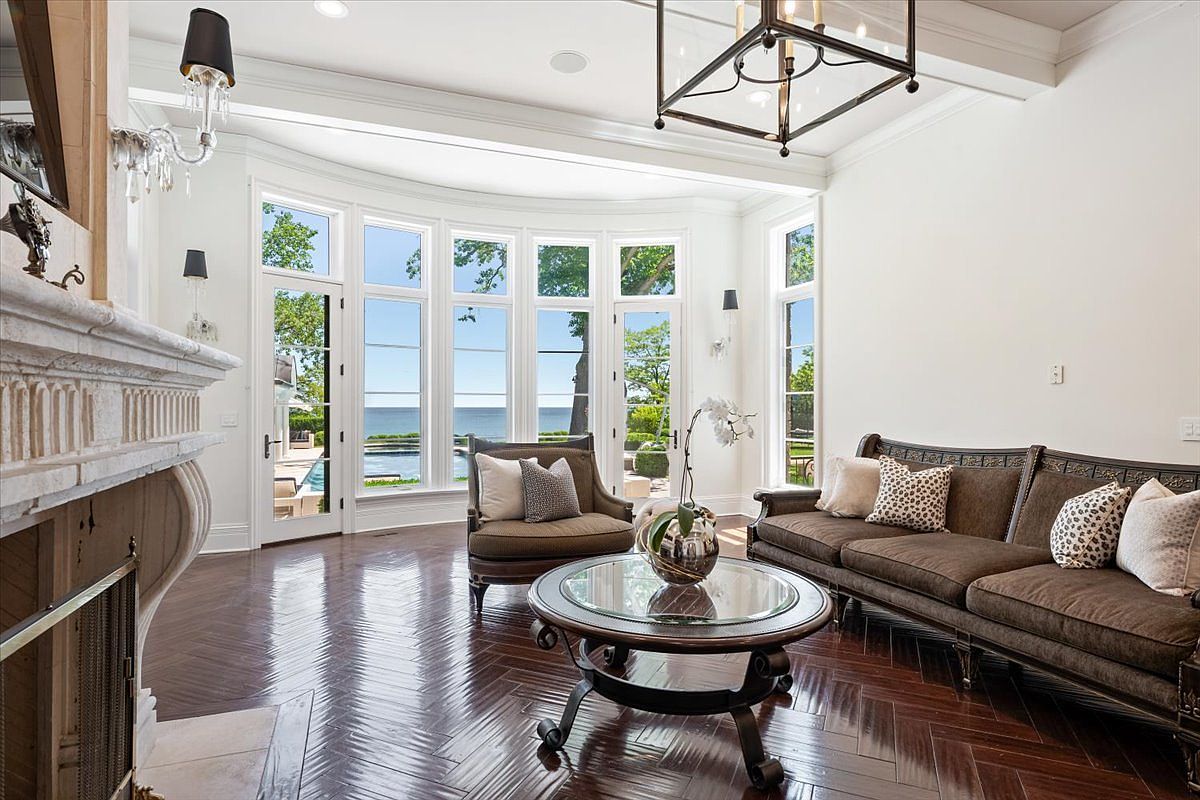 This elegant living room features rich, dark-stained wood flooring laid in a herringbone pattern and a sophisticated neutral color palette. A curved wall of tall windows provides expansive views of the outdoor pool and waterfront, while a classic stone fireplace surround adds a focal point to the space. The room is furnished with a plush brown sofa and matching armchair, centered around a circular glass-topped coffee table under a large, dark metal chandelier.