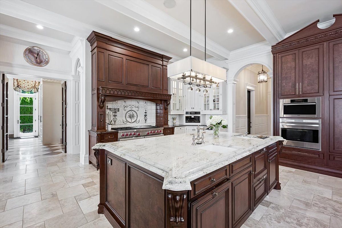This elegant kitchen features dark wood cabinetry, a large central island with a light-colored stone countertop, and a professional-grade range set beneath a decorative hood. The space is illuminated by a prominent rectangular chandelier and recessed lighting, while the light-toned tile flooring creates a bright contrast against the rich wood tones. The perspective captures the expansive layout, highlighting the seamless transition between the main cooking area and the adjacent hallway.