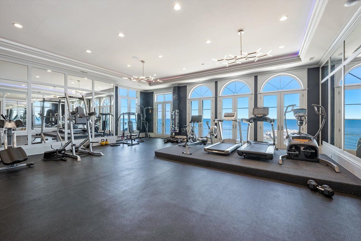This expansive home gym features a dark rubberized floor and a raised platform housing various cardio equipment, including treadmills and an elliptical. Large windows with arched tops provide panoramic views of the water, filling the room with natural light. The space is accented by modern, multi-armed light fixtures and mirrored walls that enhance the sense of scale and luxury.