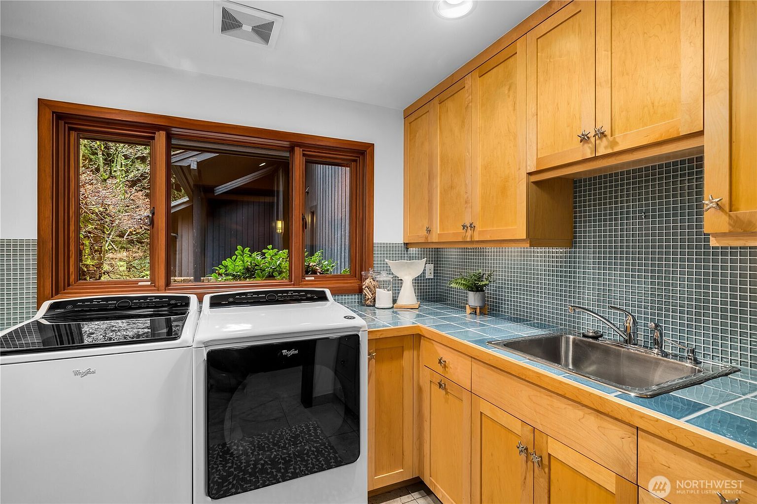 This functional laundry room features a clean, bright aesthetic with light wood cabinetry and a contrasting blue tiled countertop and backsplash. The space is equipped with a modern white Whirlpool washer and dryer set, positioned conveniently next to a stainless steel utility sink. A large window above the appliances provides natural light and a view of the exterior, creating an inviting and practical workspace.