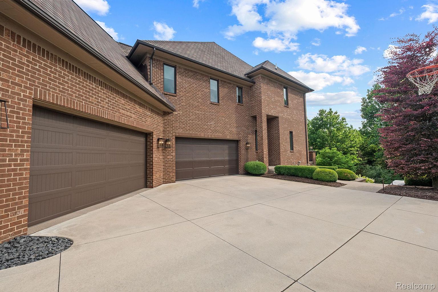 This is a front exterior view of a large, two-story brick house with a multi-car garage. The driveway is concrete and leads up to the garages. Landscaping includes trimmed bushes and a tree with reddish-purple leaves, adding to the property's curb appeal.