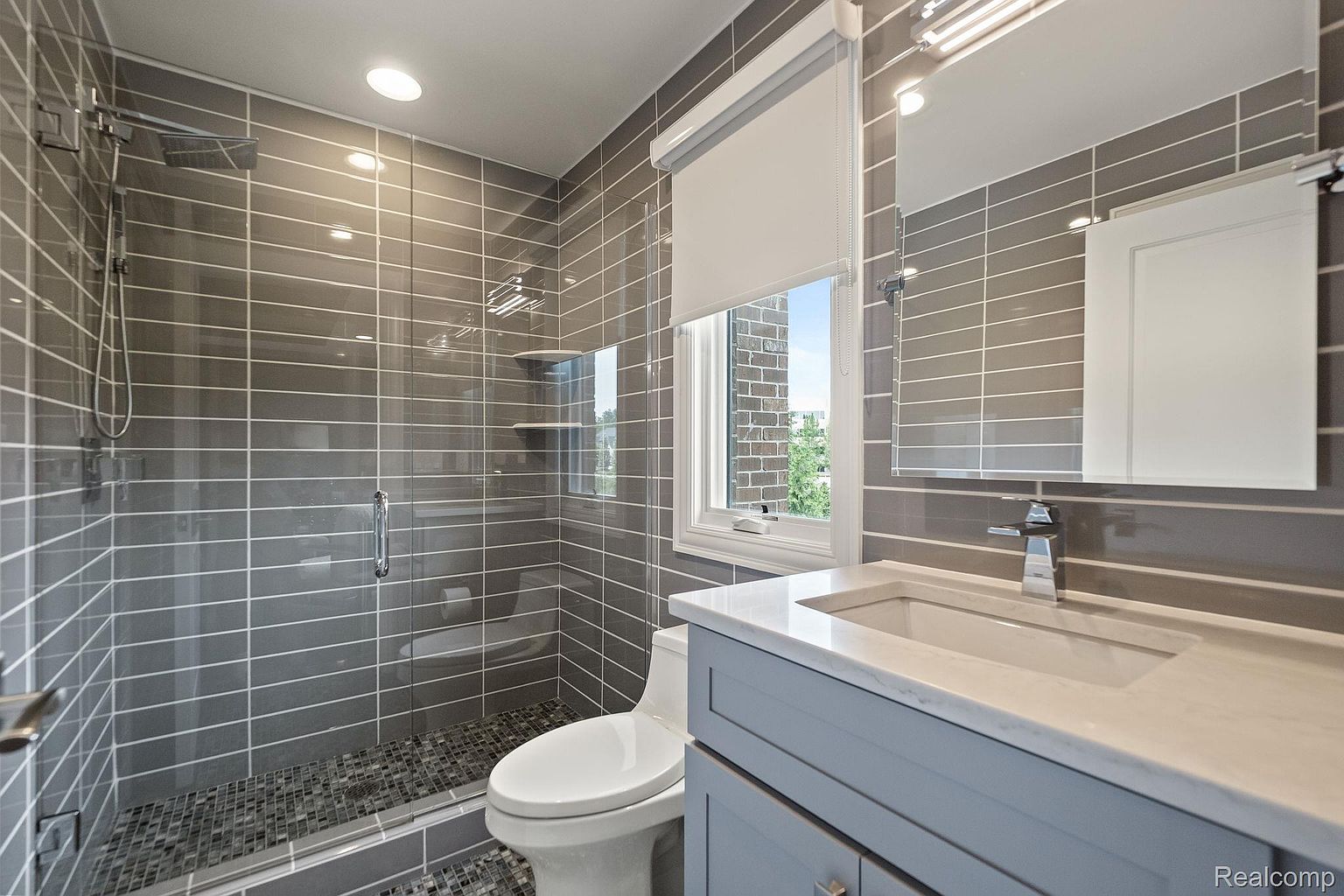 This is a modern bathroom featuring gray horizontal tiling, a glass-enclosed shower with a rainfall showerhead, and a vanity with a light blue cabinet and white countertop. The bathroom also includes a toilet and a window with a white roller shade, providing natural light. The overall impression is clean and contemporary.