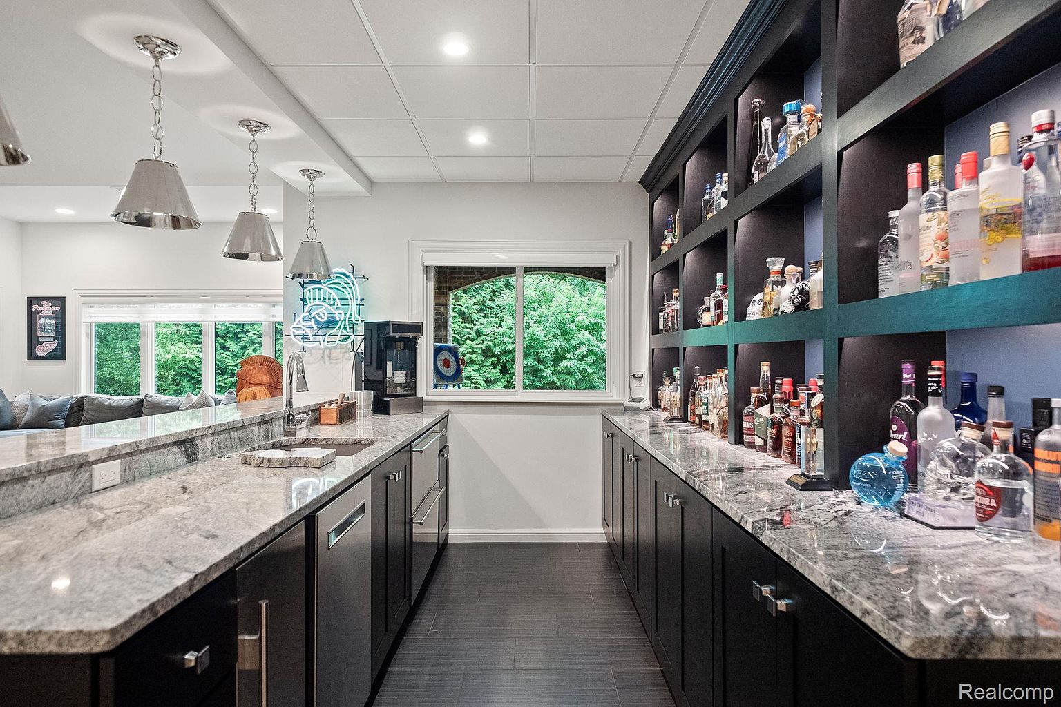 This is an interior shot of a basement bar area, featuring dark cabinetry with a light granite countertop. The bar is well-stocked with liquor bottles displayed on shelves, and there is a sink and dishwasher on the left side. A window provides natural light and a view of the greenery outside.
