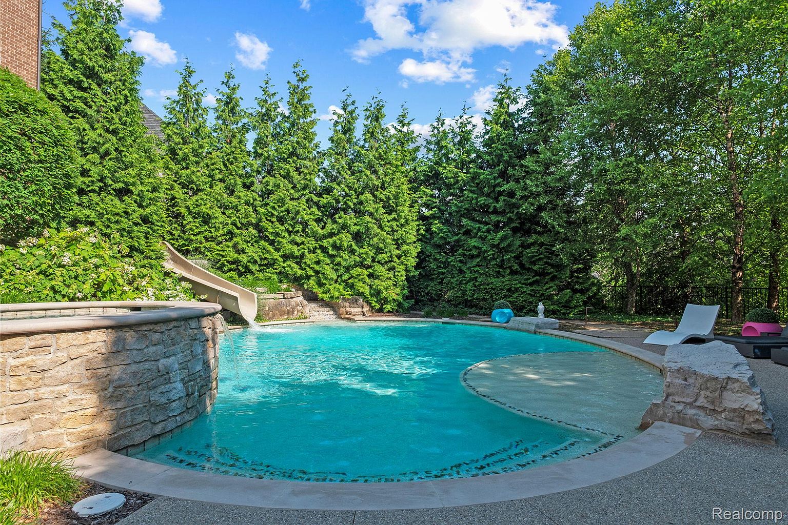 This image showcases a luxurious backyard pool area, complete with a slide integrated into a stone feature. The pool is surrounded by lush greenery, including tall evergreen trees, providing privacy and a serene atmosphere. A stone patio area with lounge chairs offers a relaxing space for sunbathing and enjoying the pool.
