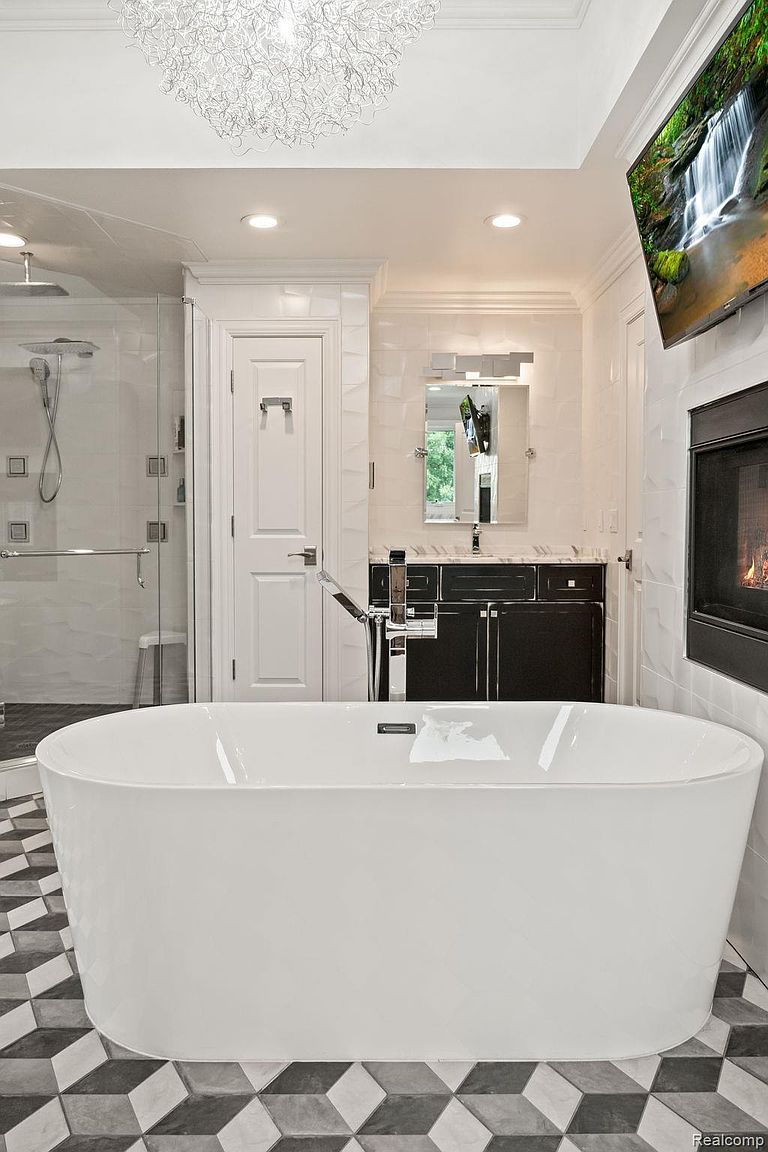 This is a luxurious primary bathroom featuring a freestanding white bathtub as the focal point, set against a backdrop of geometric patterned flooring. The room includes a glass-enclosed shower, a dark vanity with a mirror, and a fireplace with a mounted television above it, creating a spa-like atmosphere. The perspective is from a medium shot, emphasizing the depth and high-end finishes of the space.