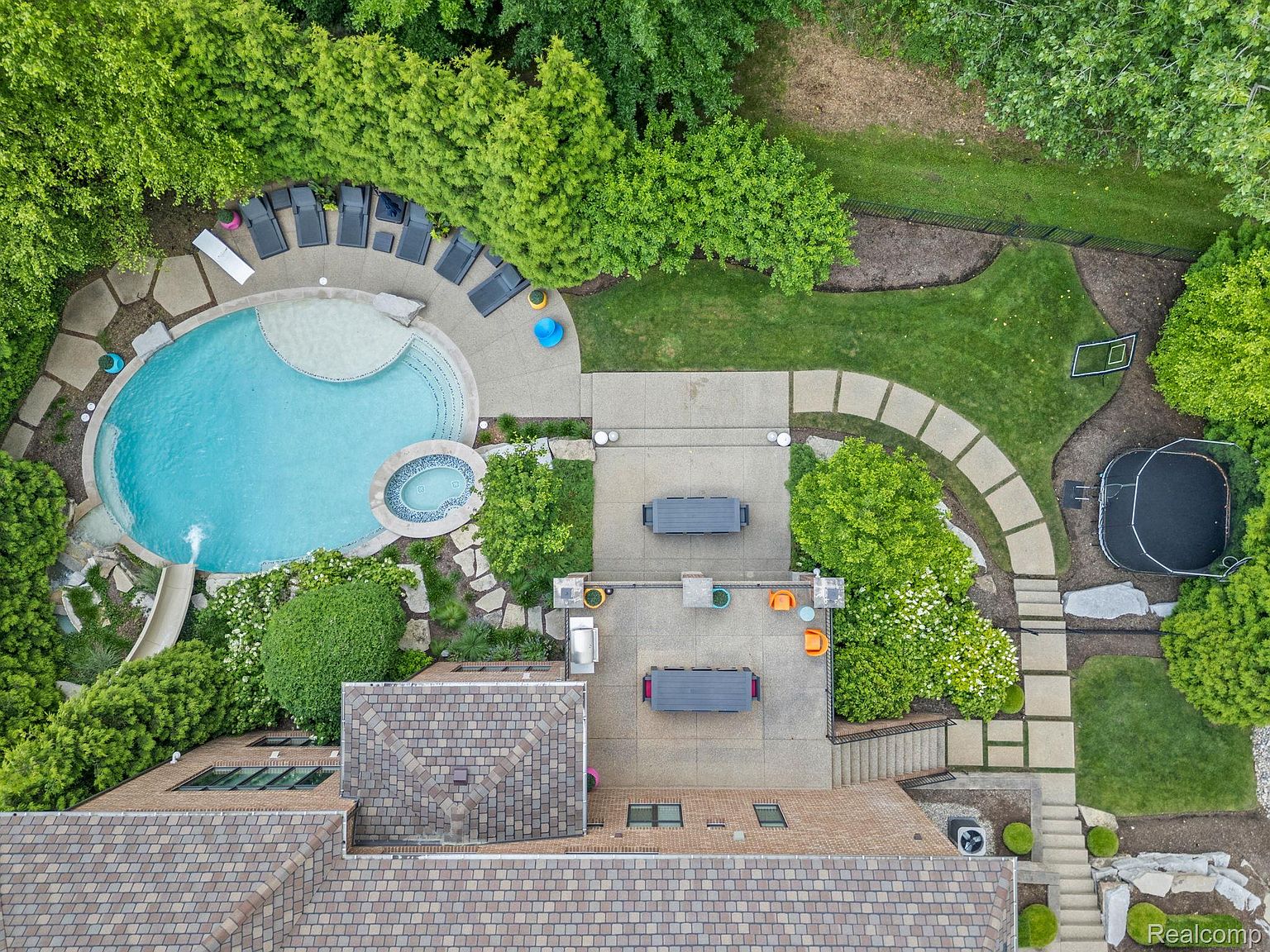 This aerial view showcases a luxurious backyard featuring a large, freeform pool with a slide and spa, surrounded by lush landscaping and patio areas. The property includes multiple outdoor dining spaces, a trampoline, and a well-maintained lawn, creating an inviting and private oasis. The roof is a mix of brown and gray tiles.