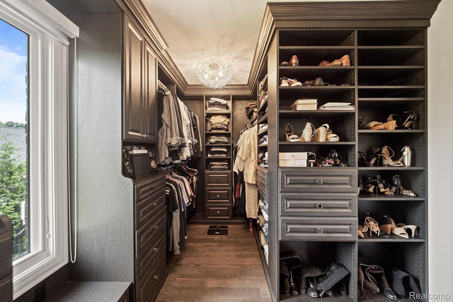 This is a well-organized walk-in closet featuring dark wood cabinetry and shelving. The closet is filled with clothing, shoes, and accessories, all neatly arranged. A decorative chandelier provides ample lighting, and the hardwood flooring adds a touch of elegance, creating a luxurious and functional space.