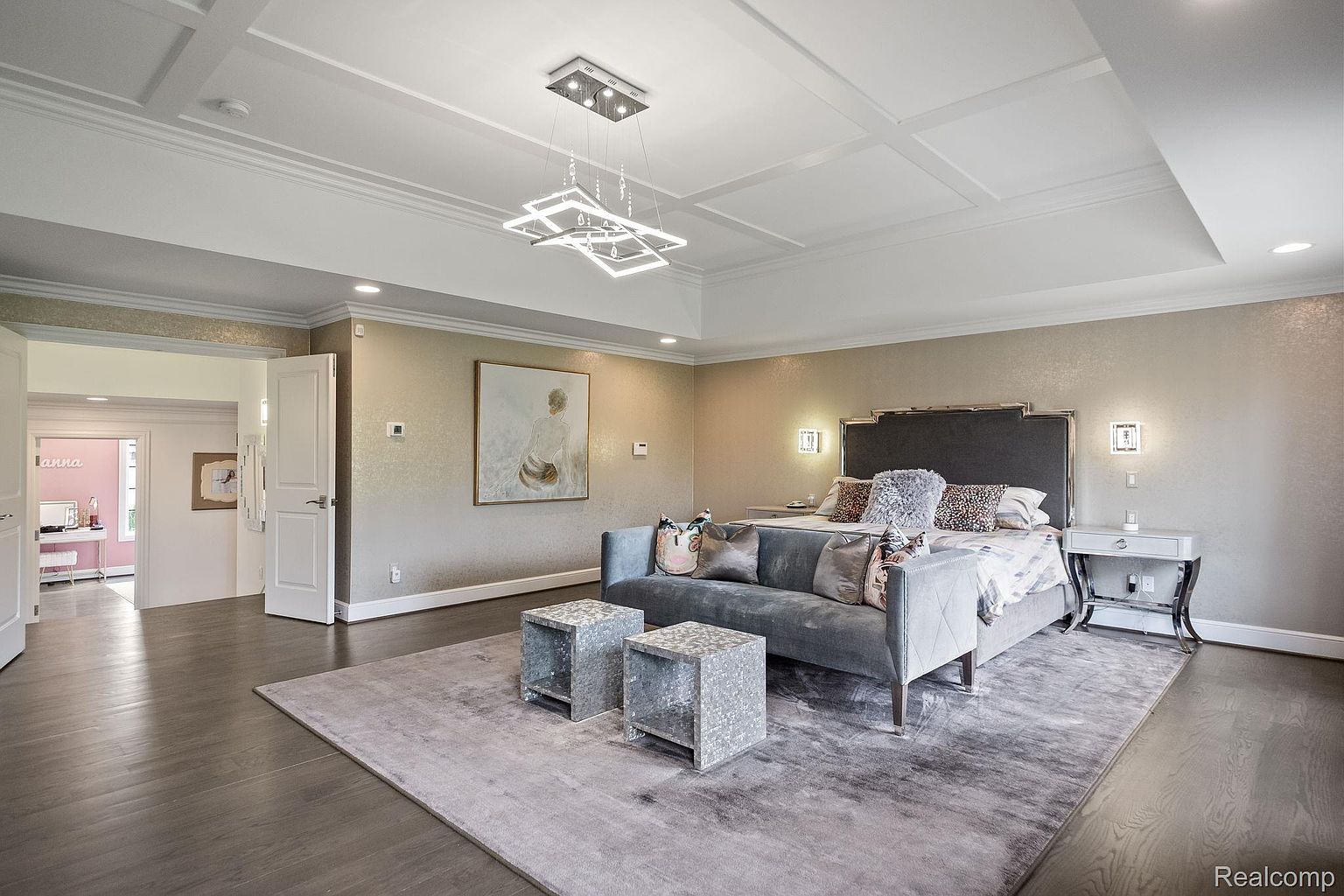 This is a primary bedroom featuring a large bed with a gray headboard and a seating area in front of it. The room has a neutral color palette with metallic wallpaper and a modern chandelier. There is a doorway leading to another room, and the overall impression is luxurious and spacious.