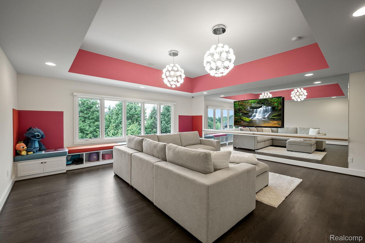 This is an interior shot of a modern living room featuring a large sectional sofa, dark hardwood floors, and a wall-sized mirror reflecting the room. The room is brightly lit with natural light from large windows and modern pendant lighting, and there is a built-in window seat with storage and decorative red accents on the walls and ceiling. A large television is mounted on the mirrored wall.