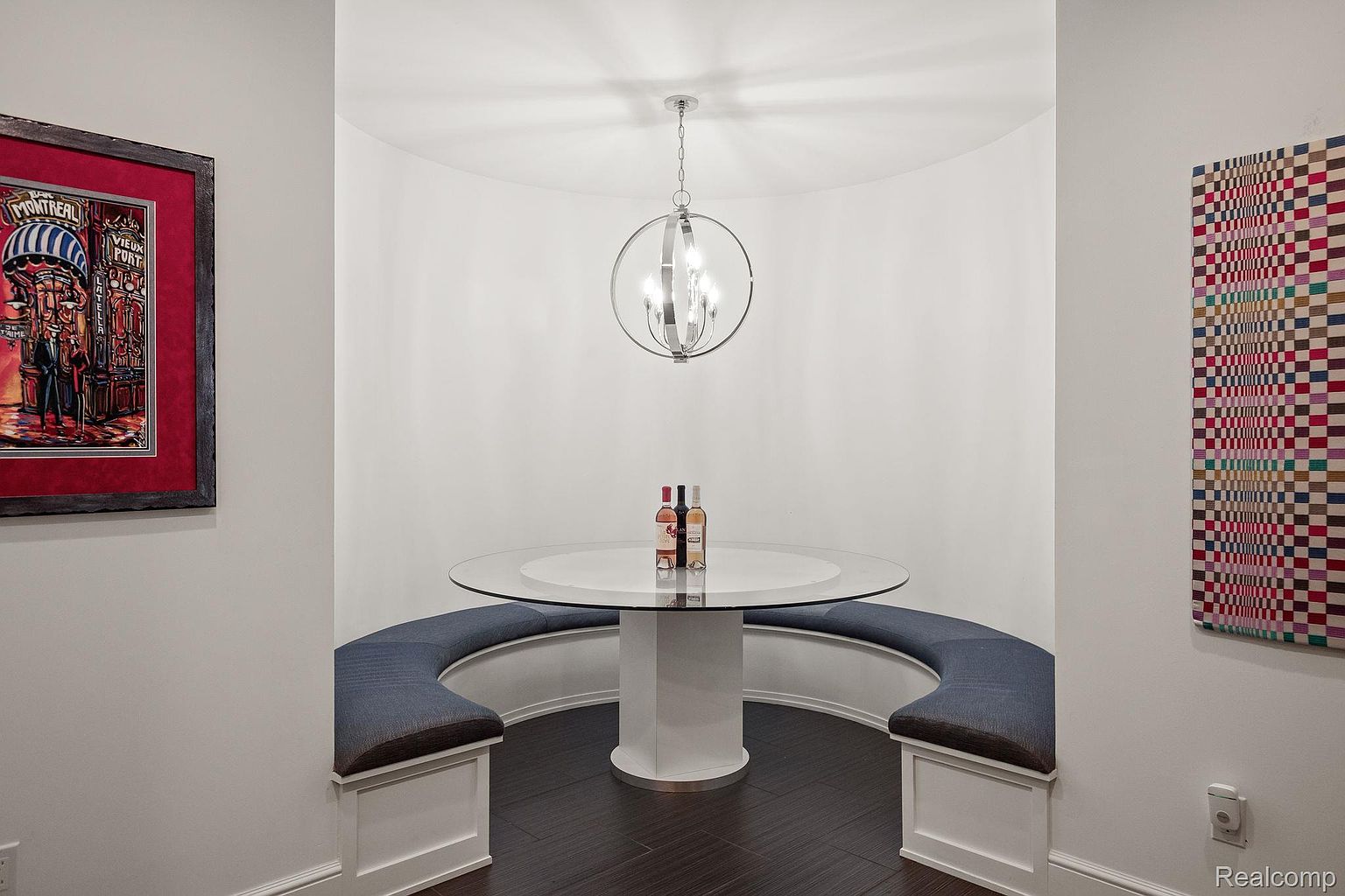 This image showcases a unique dining nook featuring a round glass-top table with a white pedestal base, surrounded by a curved banquette seating area with blue cushions. A modern chandelier hangs above the table, and artwork adorns the walls, creating a stylish and inviting space. The dark wood flooring adds a touch of elegance to the room.