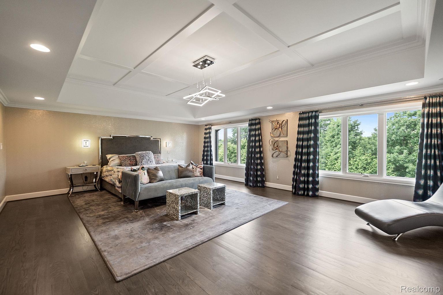 This is a spacious primary bedroom featuring a large bed with a gray headboard, a gray sofa, and two decorative side tables. The room has a tray ceiling with modern lighting, large windows offering natural light, and dark hardwood floors. The overall impression is luxurious and serene.