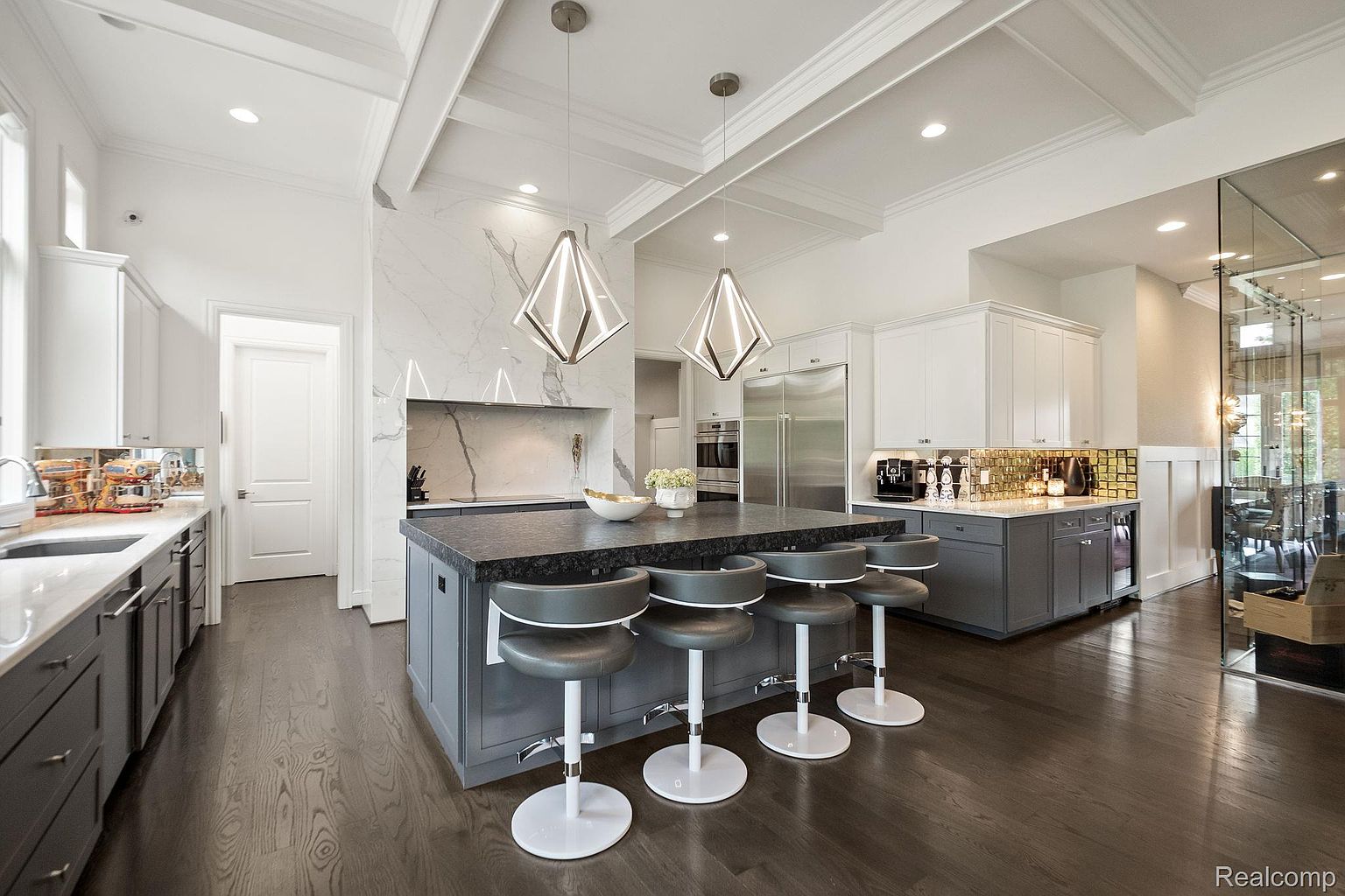 This is a bright and modern kitchen featuring a large center island with seating, dark countertops, and gray cabinetry. The kitchen also includes stainless steel appliances, white upper cabinets, and unique pendant lighting. The dark hardwood floors add warmth and contrast to the space.
